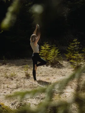 Woman in tree pose during yoga on sunny forest clearing surrounded by young spruces – outdoor yoga experience at eriro Alpine Hide retreat