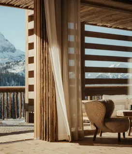 eriro Alpine Hide – Sunlit transition to wooden balcony with view of majestic mountain summit and cozy bouclé armchairs in warm light