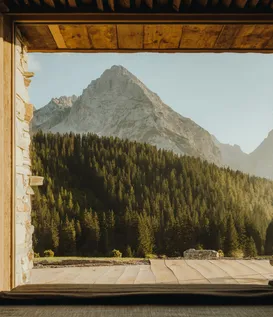 Deep-reveal stone window at eriro with opened wood slat shutter framing Wetterstein summits above larch-spruce forest, upholstered window seat