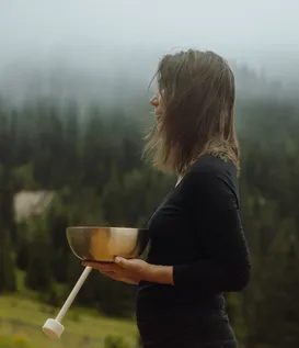 Woman holding golden singing bowl before misty alpine forest backdrop – sound therapy and mindfulness practice at eriro Alpine Hide retreat