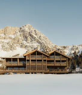 eriro Alpine Hide – Imposing wooden facade of luxury chalet with wraparound balconies before dramatic rock wall and pristine snow surface in Alps