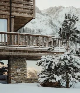 Winter exterior view of eriro Alpine Hide with wooden facade, natural stone wall, wooden balcony and snow-covered conifer before mountain panorama