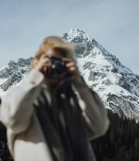 Woman in cream wool coat photographing with camera – behind her snow-covered rock summits of Wetterstein rising into clear winter sky