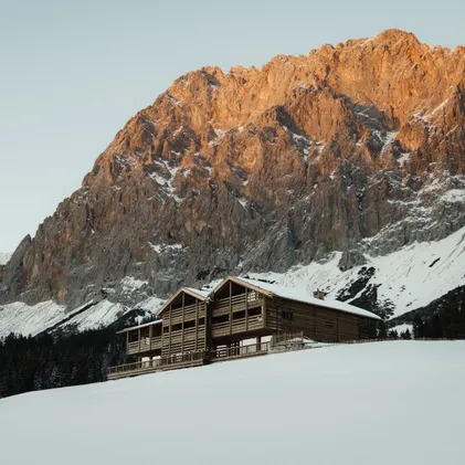 eriro Alpine Hide wooden building in snow-covered winter landscape before imposing Zugspitze rock wall in golden alpenglow at sunset