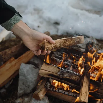 Hand placing rough birch log into crackling campfire at Wetterstein – orange flames, glowing embers and sparks before snow-covered ground