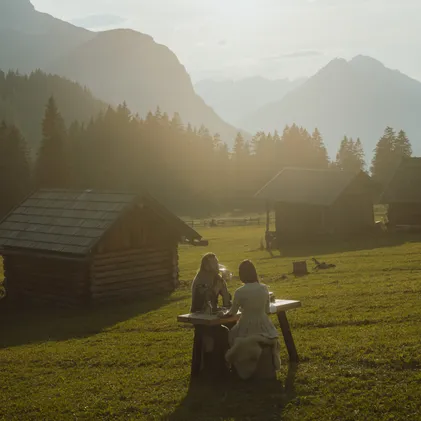 Golden hour on Tyrolean alp: Couple enjoying dinner at wooden table between historic hay barns before Wetterstein panorama at eriro