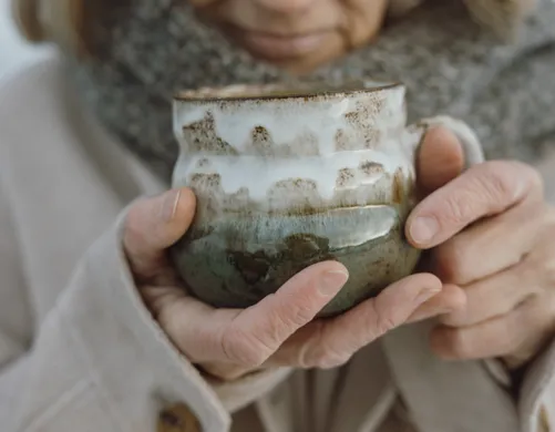 Hands cradling hand-thrown ceramic cup with gradient glaze in white and greenish-blue, wool scarf – warming tea break at eriro Zugspitze resort