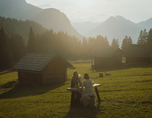 Golden hour on Tyrolean alp: Couple enjoying dinner at wooden table between historic hay barns before Wetterstein panorama at eriro