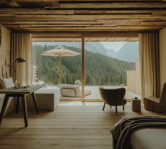 Eriro Suite living area with reclaimed wood ceiling, textured wool wall, sheepskin armchair before panorama window with terrace and view of conifer forests and mountain summits