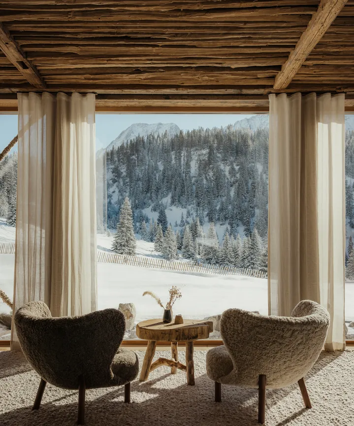 eriro Alpine Hide – Cozy bouclé armchairs before panorama window with breathtaking view of snow-covered alpine valley and frosted conifer forests